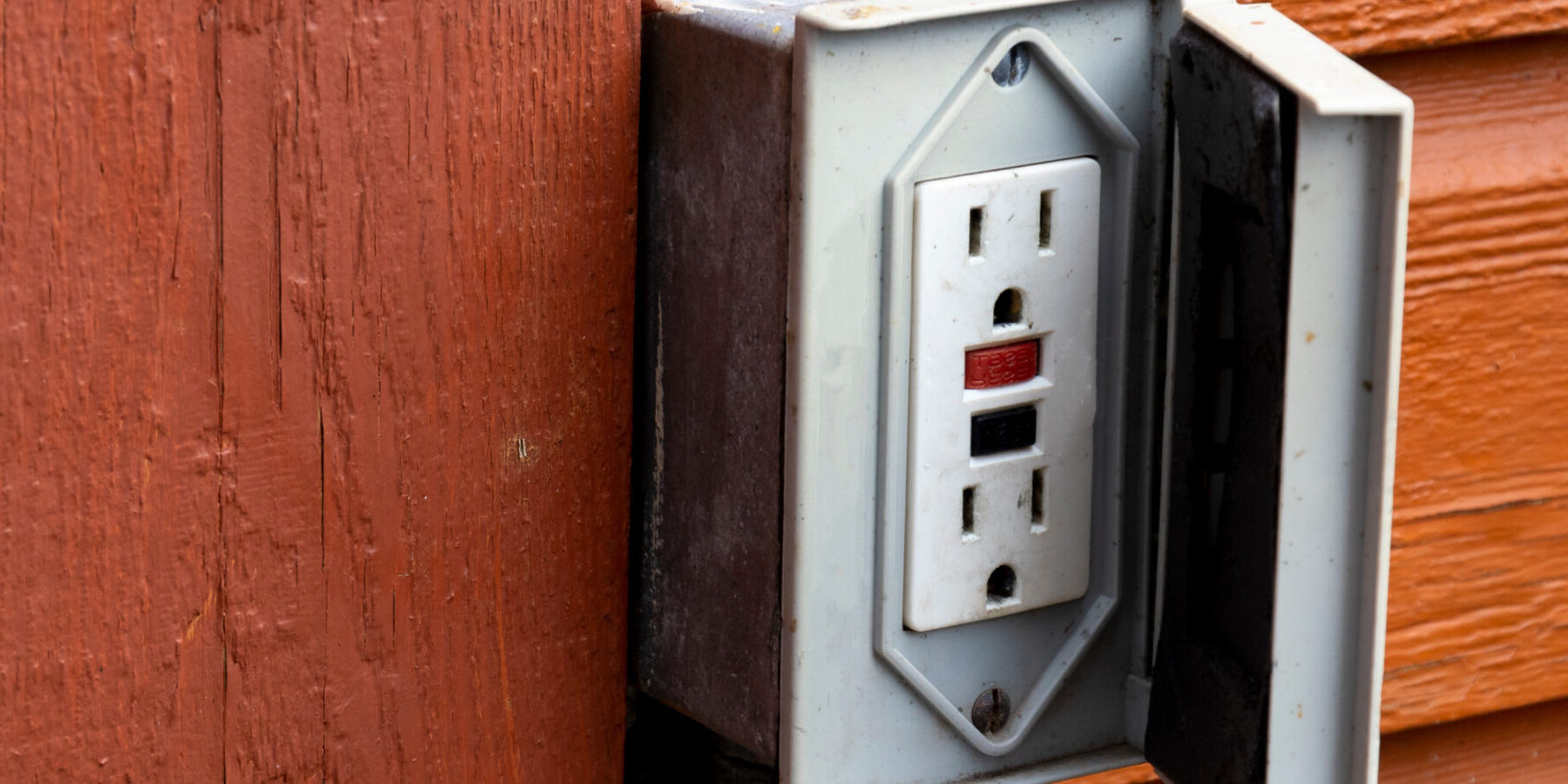 An image of an old outdoor electrical outlet on the exterior wall of a reddish brown building.