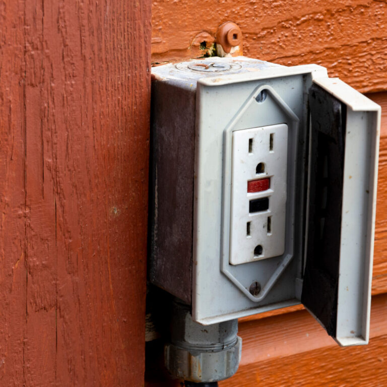 An image of an old outdoor electrical outlet on the exterior wall of a reddish brown building.